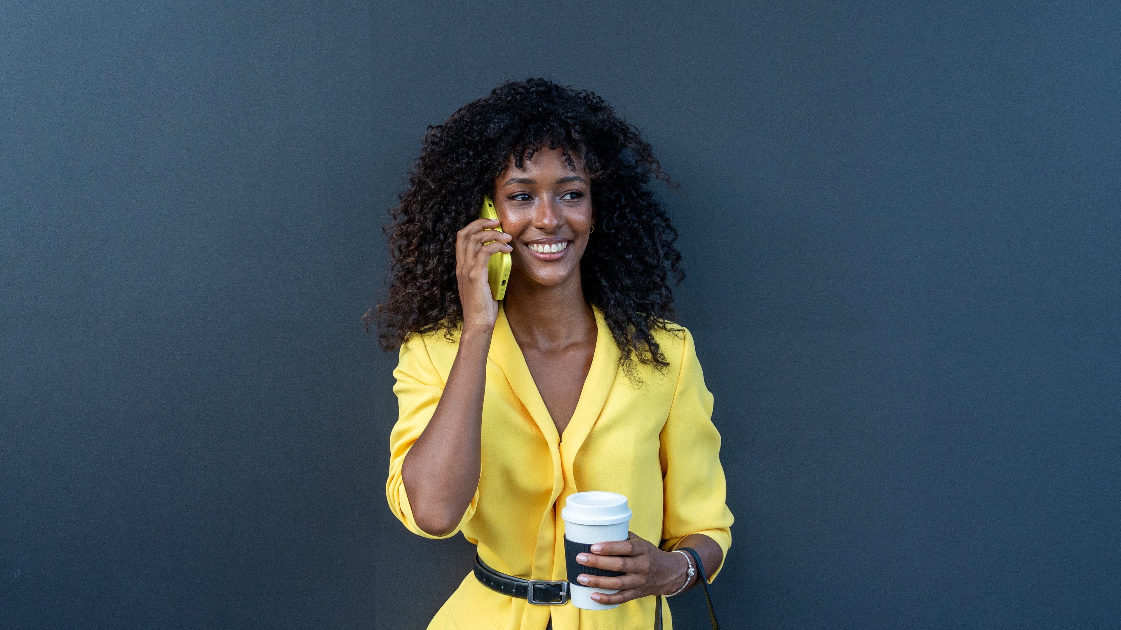 Woman in yellow jacket holding a phone and a coffee cup against a dark background